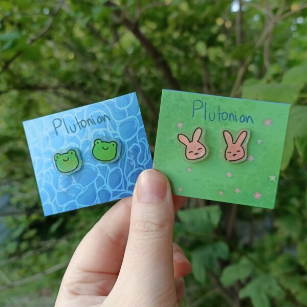 A white feminine hand holding two backing cards with two lots of studs, one design being a frog, the other a bunny