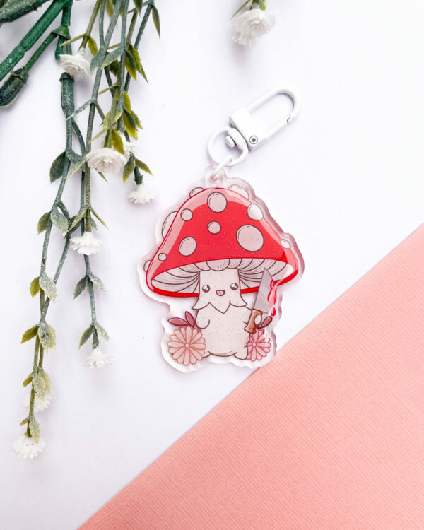 An acrylic keychain depicting a cute red cap mushroom holding a knife, smiling innocently, surrounded by pink flowers. It is placed against a white and red background.