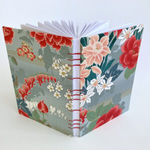 An A5 sized notebook featuring red, white, and orange flowers against a grey background. The book is upright and opened.