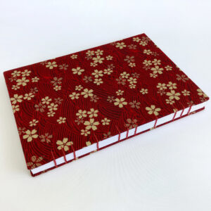 An A5 sized notebook featuring small gold cherry blossoms against a dark red background with faint wave patterns. The book is set at an angle.
