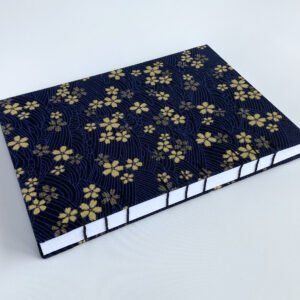 An A5 sized notebook featuring small gold cherry blossoms against a dark blue background with faint wave patterns. The book is set at an angle.