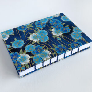 An A6 sized notebook featuring blue blossoms against a blue background with gold curved lines. The book is set at an angle.