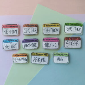 a series of 11 shrink plastic pins depicting various pronouns on different coloured paper. The pins each read "my pronouns are" in a colourful header with the selected pronouns under that in a white area. The pronouns are he/him