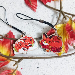 Photo of two wooden charms with braided phone straps. The first design is a pair of foxes huddled together in snow. The second design is a single fox curled up on a pile of autumn leaves.