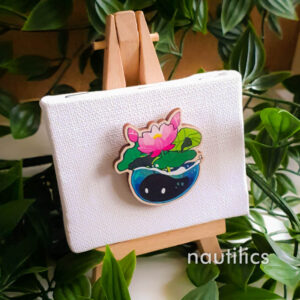 Photo of a wooden pin displayed on a mini canvas and easel, surrounded by decorative leaves. The pin is of a small black blob-shaped creature with simplified cute eyes, hiding underwater with lotus flowers and lotus pads blooming above it.