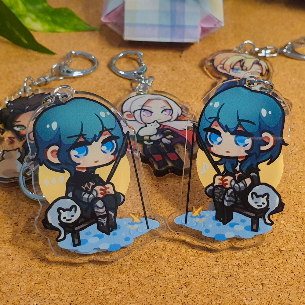 Fire Emblem: Three Houses Acrylic Charms by Nautilics Photo of acrylic charms of characters from Fire Emblem: Three Houses. The main focus of this photo are two Byleths (both the F and M designs) fishing next to a small cat, but other characters are visible in the background.