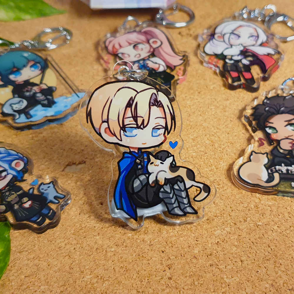 Fire Emblem: Three Houses Acrylic Charms by Nautilics Photo of acrylic charms of characters from Fire Emblem: Three Houses. The main focus of this photo is Dimitri petting a cat on his lap, but other characters are visible in the background.