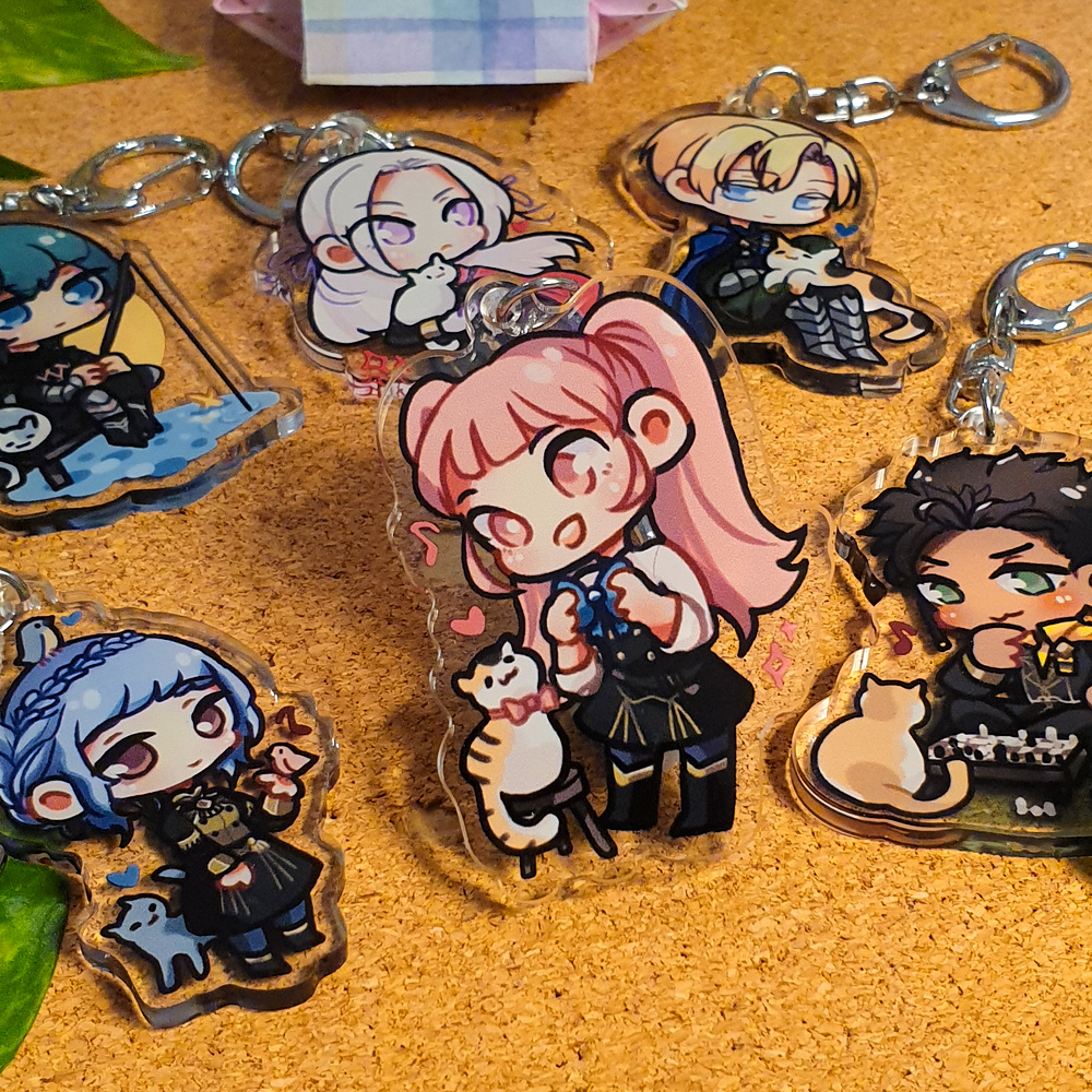 Fire Emblem: Three Houses Acrylic Charms by Nautilics Photo of acrylic charms of characters from Fire Emblem: Three Houses. The main focus of this photo is Hilda playing dressup with a cat, but other characters are visible in the background.