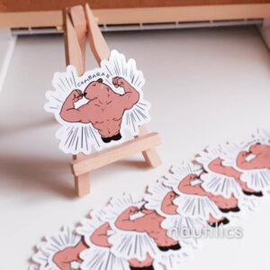 Photo of a sticker of an anthropomorphised capybara with a very buff body, flexing its arms with dramatic emphasis lines around it. The sticker is displayed on a mini easel and surrounded by even more identical stickers.